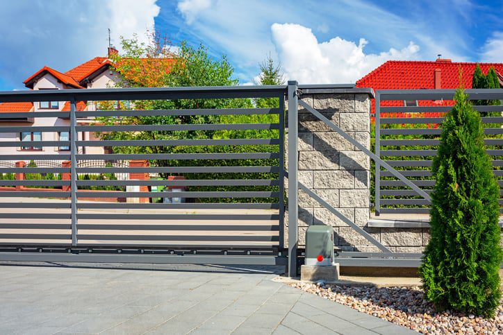 A modern horizontal metal slat gate with an automated opener is set beside a stone pillar, with red-roofed houses and trees in the background.