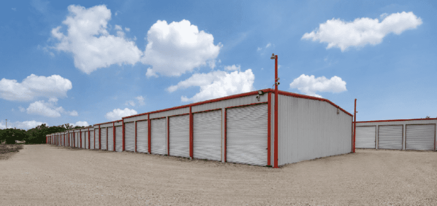 A long row of storage units with closed metal doors, surrounded by a gravel lot, under a blue sky with clouds.