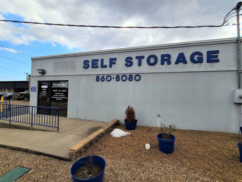 A gray building with a "Self Storage" sign and phone number 860-8080. Blue planters are placed outside on gravel. An entrance door is on the left with posted information.