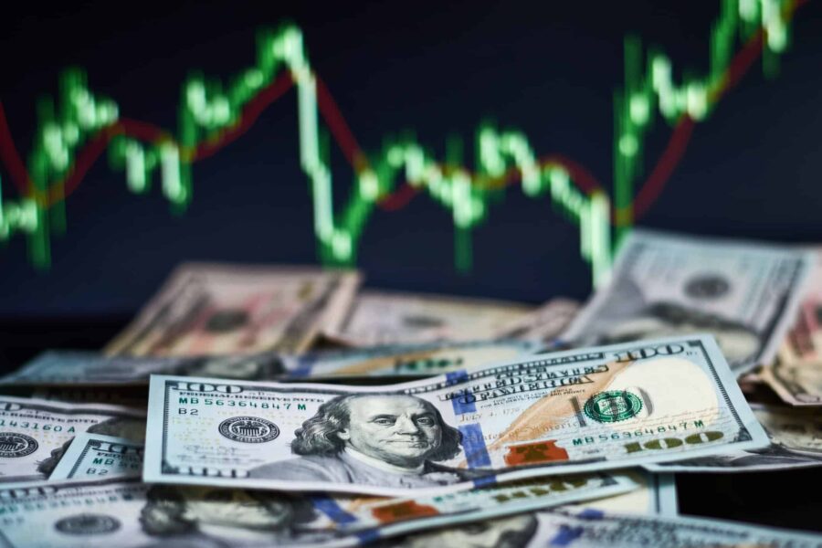 Various denominations of U.S. dollar bills spread out with a blurry stock market graph showing upward and downward trends in the background.