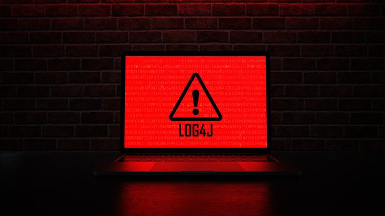 Laptop screen displaying a red warning triangle with an exclamation mark and the text "LOG4J" against a background of binary code, set against a dark brick wall backdrop.