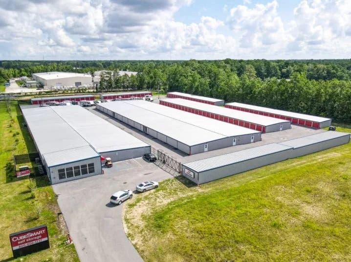 Aerial view of a storage facility with rectangular metal buildings, surrounded by greenery and a few vehicles parked in front. A sign with "CubeSmart" is visible.
