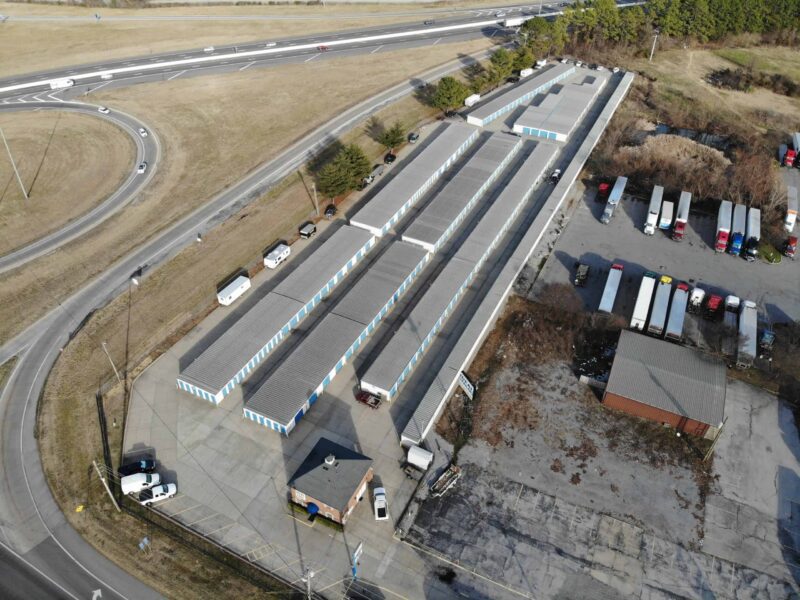 Aerial view of a self-storage facility with multiple rows of storage units, located near a highway interchange, surrounded by grass and parked vehicles.