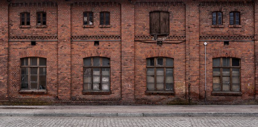 Old brick building with six large windows, some broken or boarded. A wooden double door is above the center window. A cobblestone street runs in front of the building.