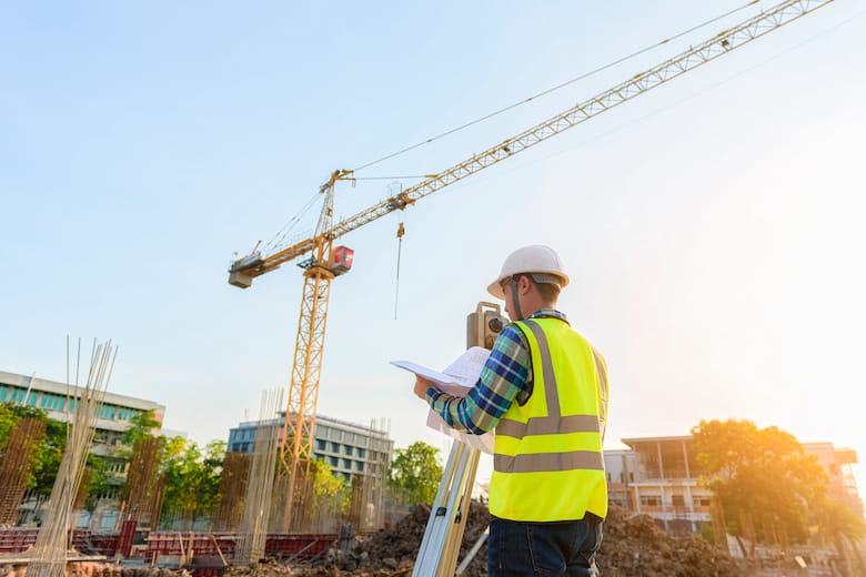 Construction worker in a safety vest and helmet reviews plans at a building site with a crane and unfinished structures in the background.