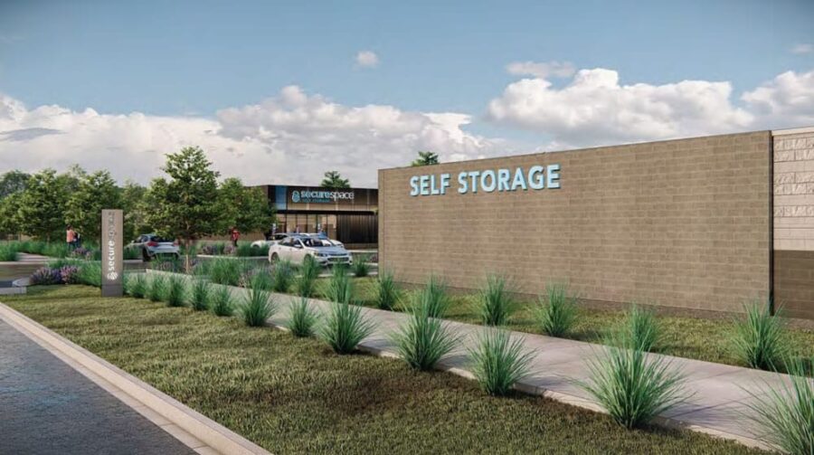 Self-storage facility with beige wall and blue lettering, surrounded by neatly landscaped greenery. Cars are parked near the building, which has a sign reading "securespace.