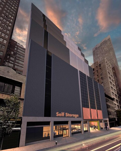 Tall, modern self-storage building with dark facade, illuminated sign, and surrounding cityscape under a pinkish clouded sky at dusk.