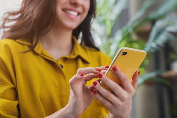 A person wearing a yellow shirt uses a yellow smartphone, smiling slightly. Green plants are blurred in the background.