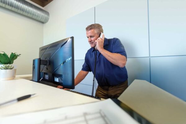 A person in a blue shirt talks on a phone while looking at a computer monitor in an office setting.