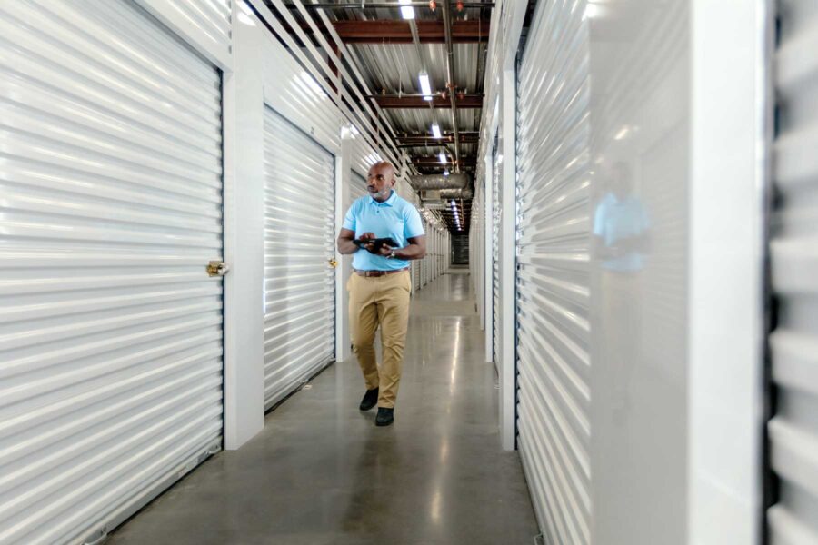 A person in a light blue shirt and beige pants walks through a storage facility hallway, holding a tablet. The corridor has closed storage units with white rolling doors.