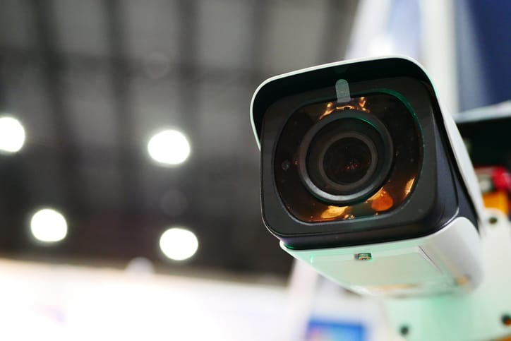 Close-up of a security camera with a wide lens, mounted and angled downwards. The blurred background shows a ceiling with bright circular lights.