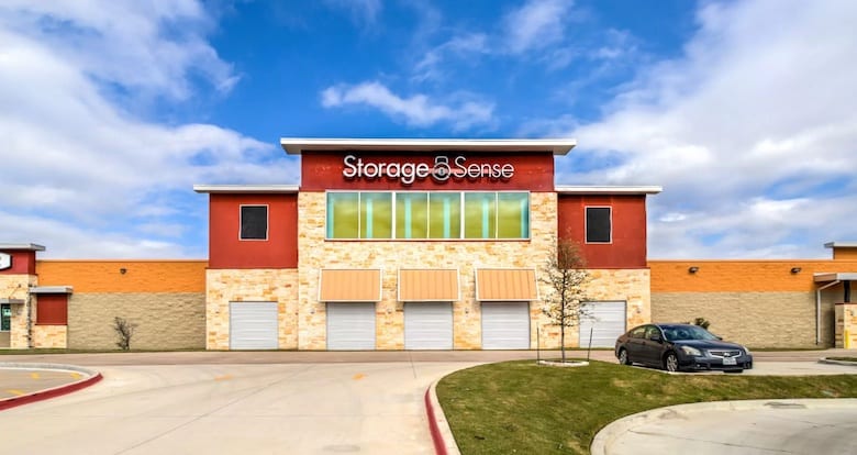 Storage facility with the sign "Storage Sense" on a two-story building. Several garage doors are visible, and a car is parked nearby. The sky is partly cloudy.