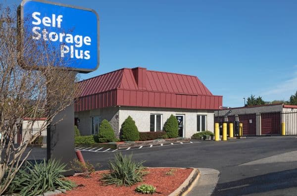 Self Storage Plus facility with a red-roofed building and a blue sign. Driveway leads to storage units behind a gate. Bushes and mulch decorate the front area.
