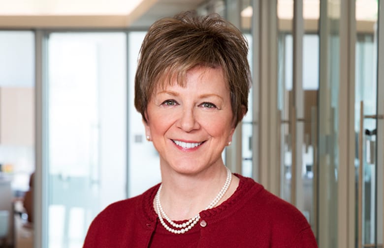 A person with short brown hair and a pearl necklace stands smiling in a modern office setting with glass walls.