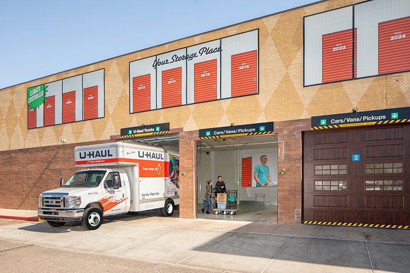 A moving truck is parked outside a storage facility. A man with a cart stands inside near open storage units. The building has orange doors and a sign reading "Your Storage Place.