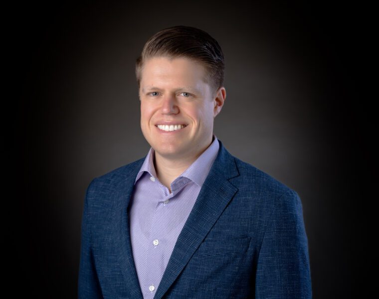 A man wearing a blue blazer and purple shirt smiles against a dark gradient background.
