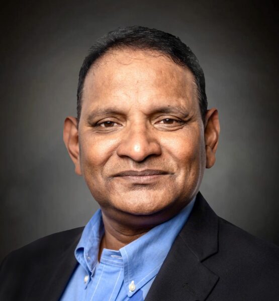 A middle-aged man in a blue shirt and dark blazer poses against a dark, neutral background, looking directly at the camera with a slight smile.