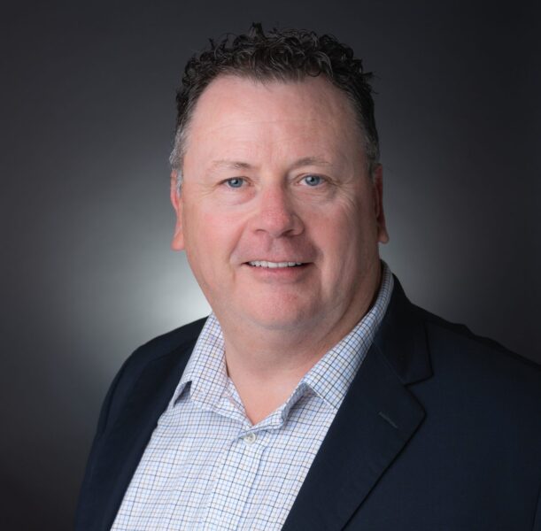 A middle-aged man with short curly hair wearing a dark blazer and a checked shirt, posing in front of a plain dark background.