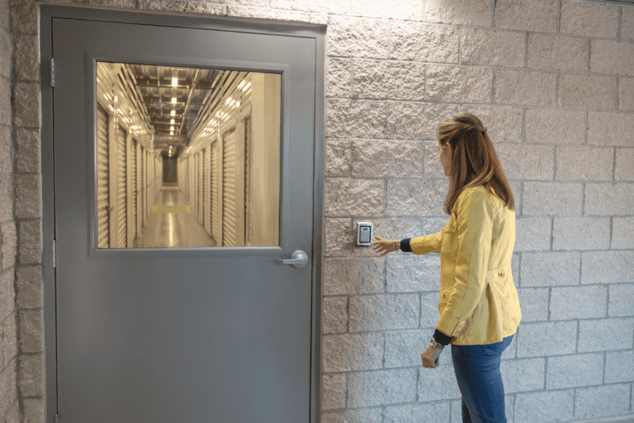 A person in a yellow jacket uses a keypad next to a grey door with a window showing a well-lit hallway.