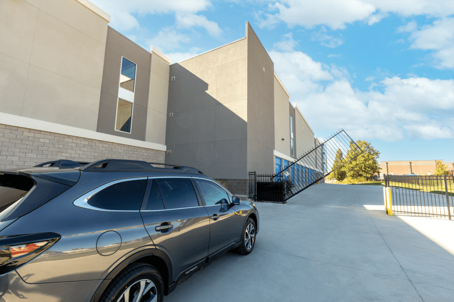 Dark SUV parked near a building with beige and gray walls. A broken fence leans against the structure. Clear blue sky with scattered clouds above.