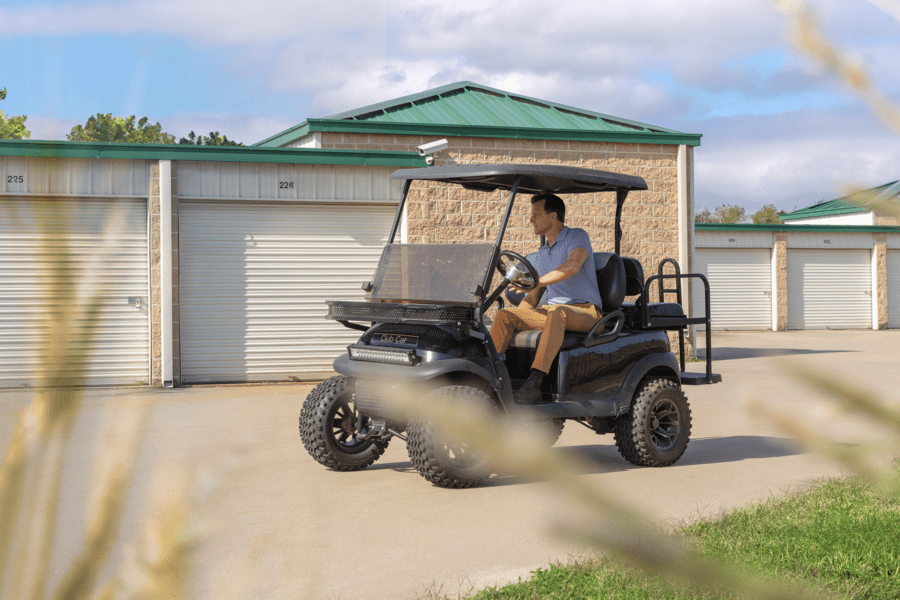 A person drives a black golf cart on a concrete path in front of storage units with green roofs, surrounded by greenery.