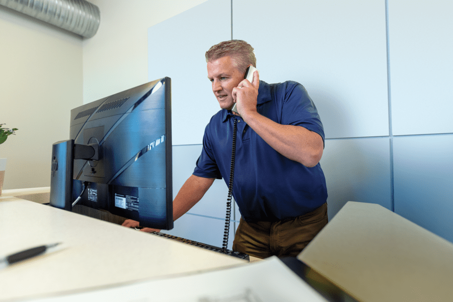 Man in a blue shirt stands at a desk, talking on a phone and looking at a computer monitor.