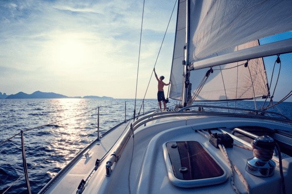 Person standing on a sailing boat, adjusting the sail, with calm ocean and distant hills under a bright sky.