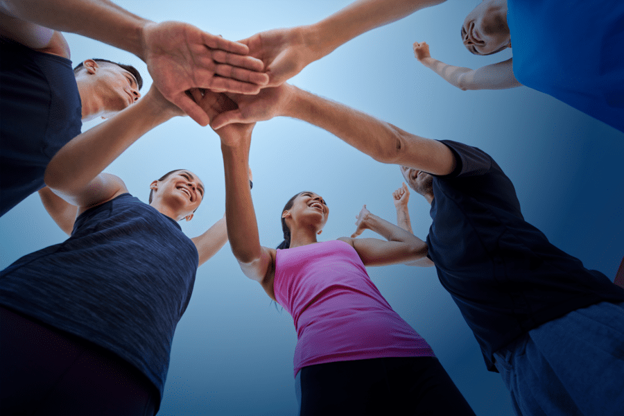 A group of people stand in a circle with their hands joined in the center, looking upwards and smiling against a blue sky background.