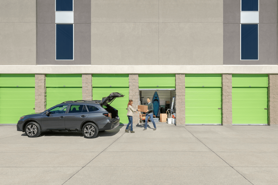 People loading a car with boxes in front of lime green storage units. Some units are open, revealing various stored items.