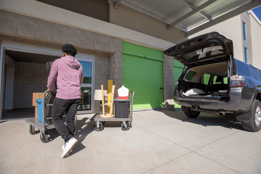 A person in a hoodie pushes a cart with boxes towards a storage unit. Another cart is nearby. An SUV with an open trunk is parked beside the unit.