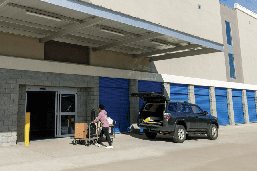 Person pushing a cart with boxes towards storage unit building with blue doors; parked SUV nearby with the trunk open.