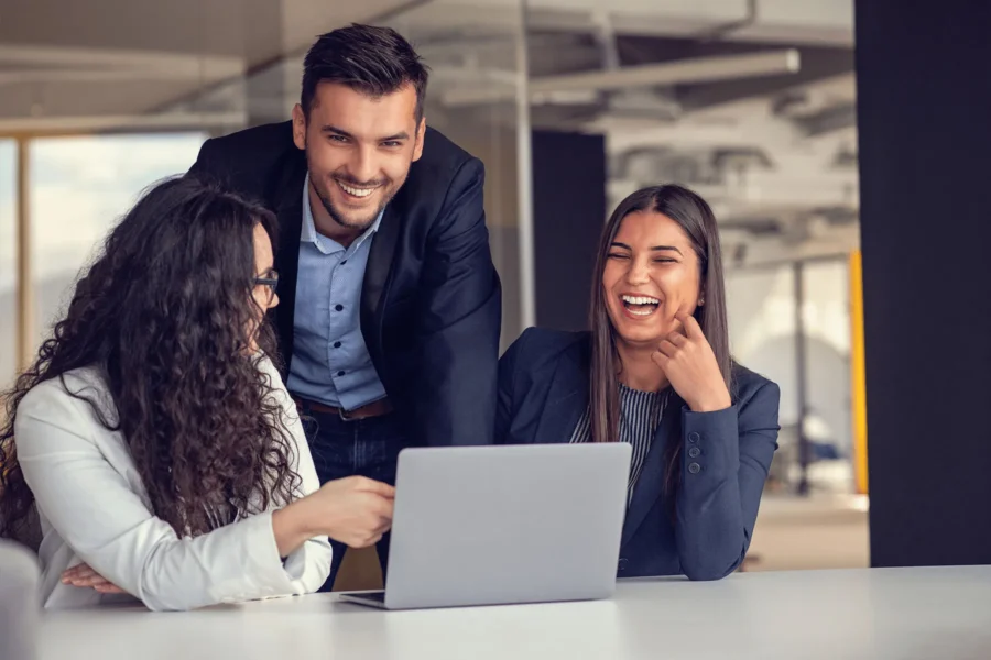 Three people in business attire are gathered around a laptop in an office setting, smiling and engaging in conversation.