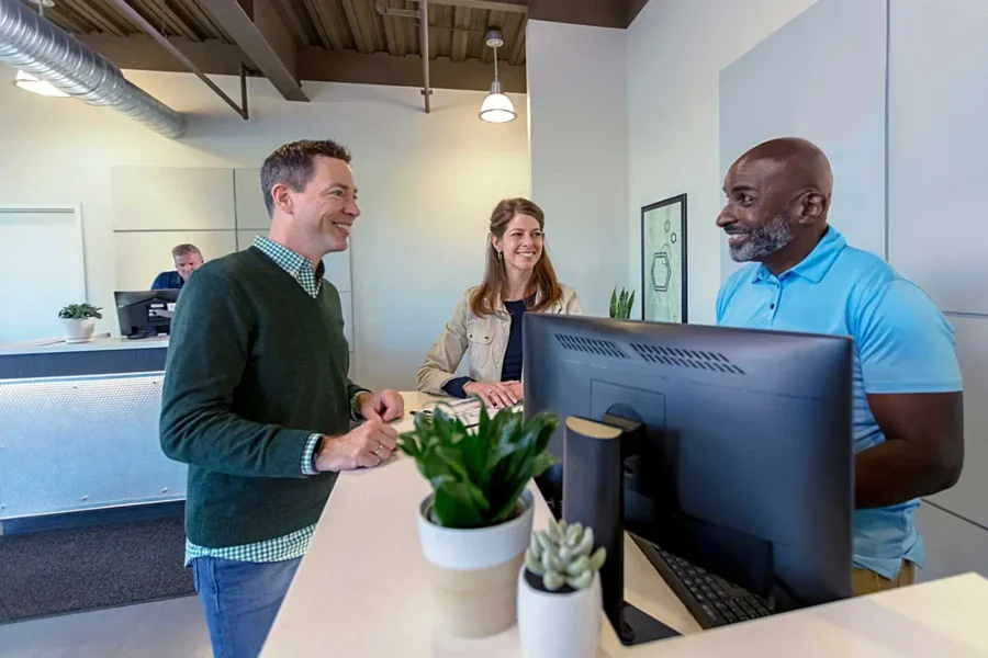 A man in a light blue polo is helping two self-storage customers at his property management computer station.