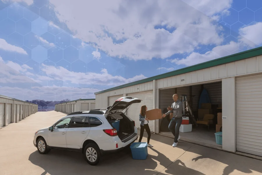 Two people are unloading boxes from a white SUV into a storage unit on a sunny day, with several storage units visible in the background.